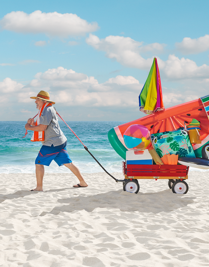 Older white man at the beach smiling and dragging behind him a wagon with various beach supplies and toys in it